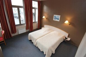 A hotel room with a double bed, two bedside lamps, a window with curtains, a chair, and a radiator. - HÔTEL LES FRANGINS (Saint-Omer)