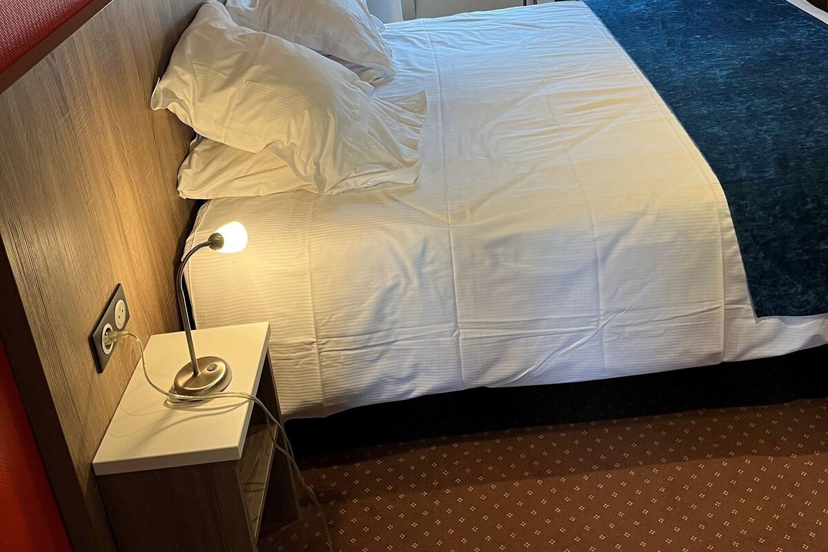 A hotel room with a bed, a bedside lamp, a window with a view of a building, and a red wall.