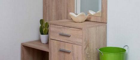 A wooden vanity with a mirror, a small shelf with flowers, a potted plant, and a green trash can.