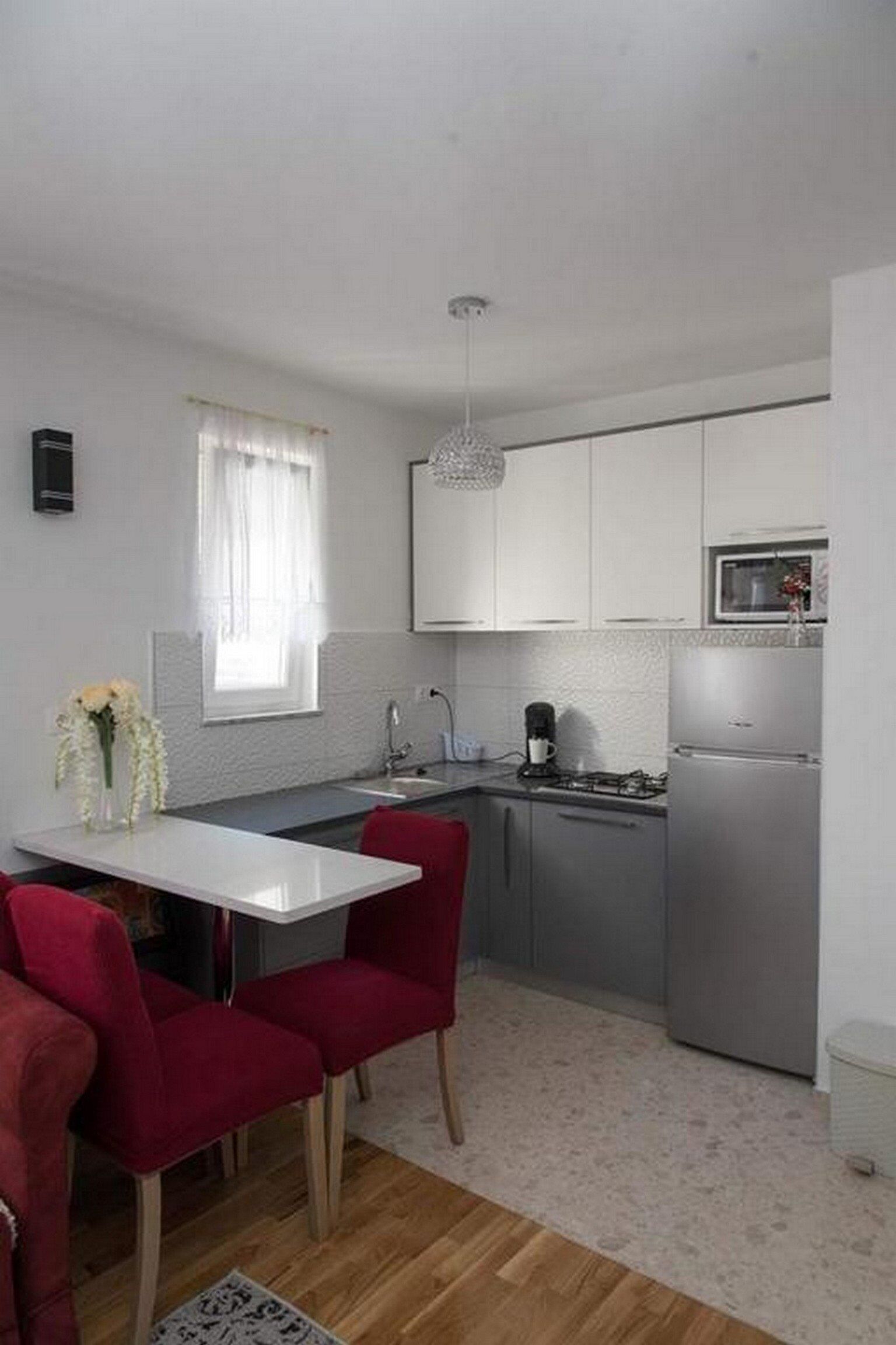 A compact kitchen with white cabinets, a stainless steel refrigerator, and a dining area with red chairs.