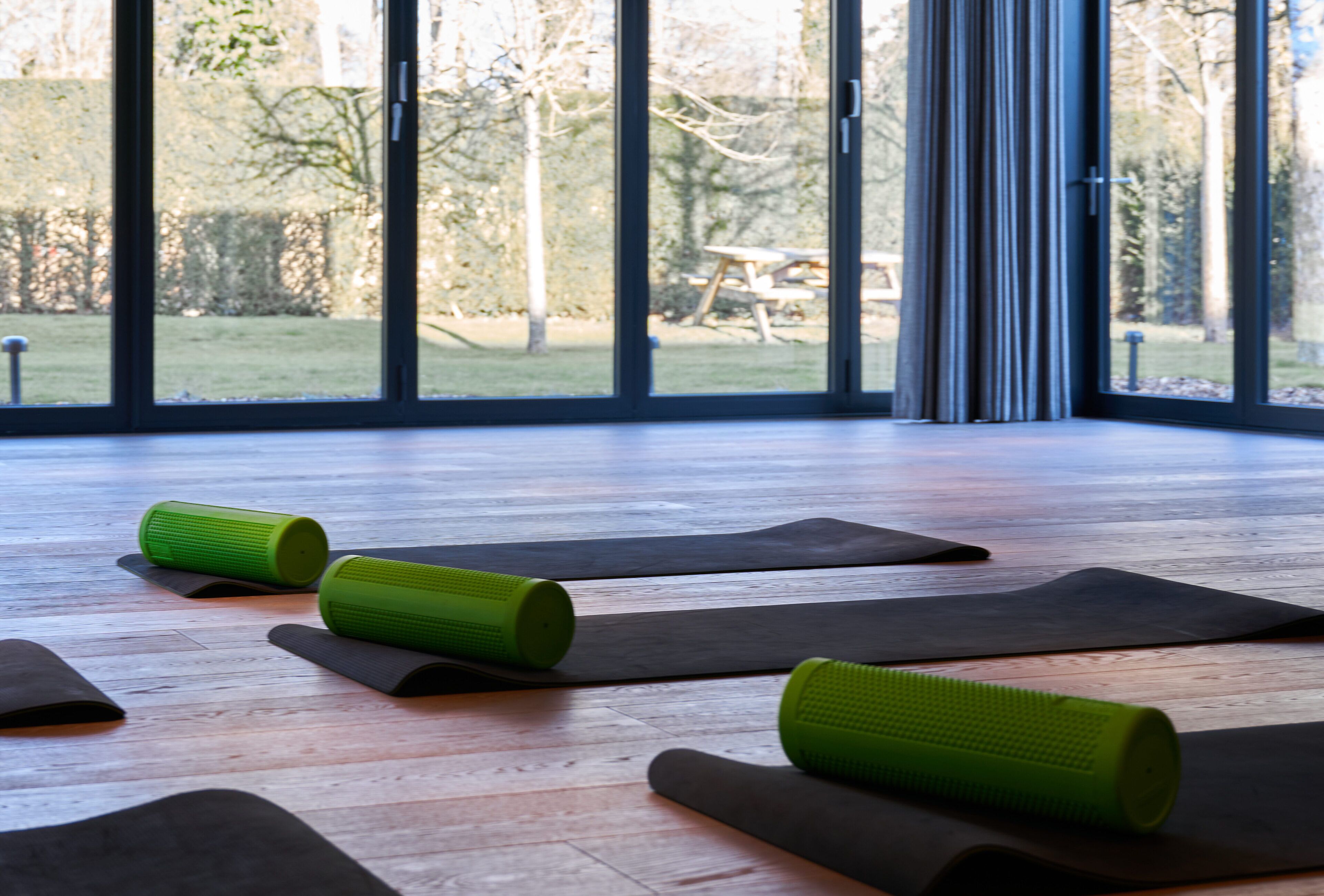 Une pièce dotée de grandes fenêtres, d’un plancher en bois et de tapis de yoga et de rouleaux en mousse disposés sur le sol.