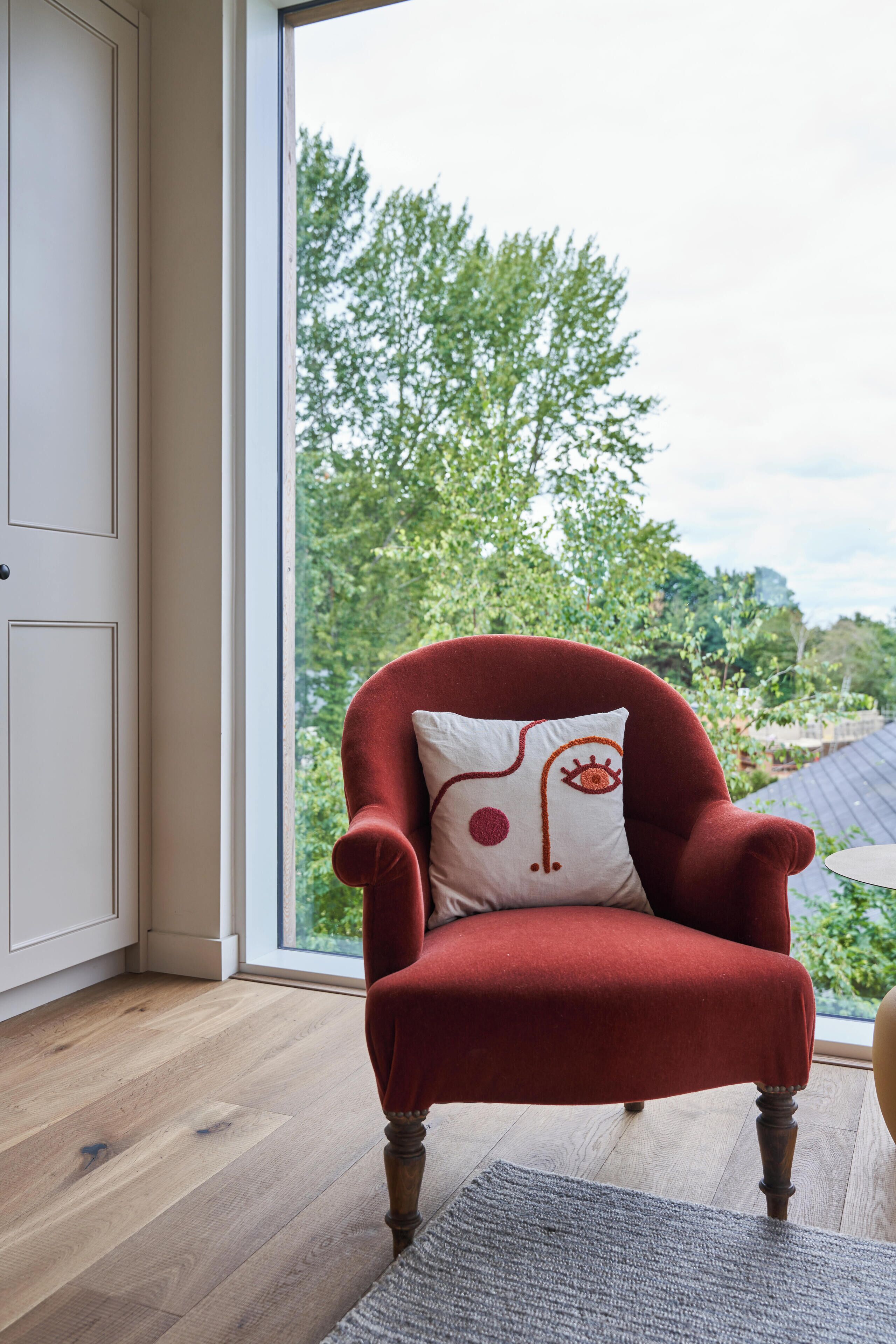 A red armchair with a white pillow featuring an abstract design, positioned in a room with wooden flooring and a view of greenery outside.