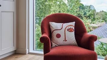 A red armchair with a white pillow featuring an abstract design, positioned in a room with wooden flooring and a view of greenery outside.
