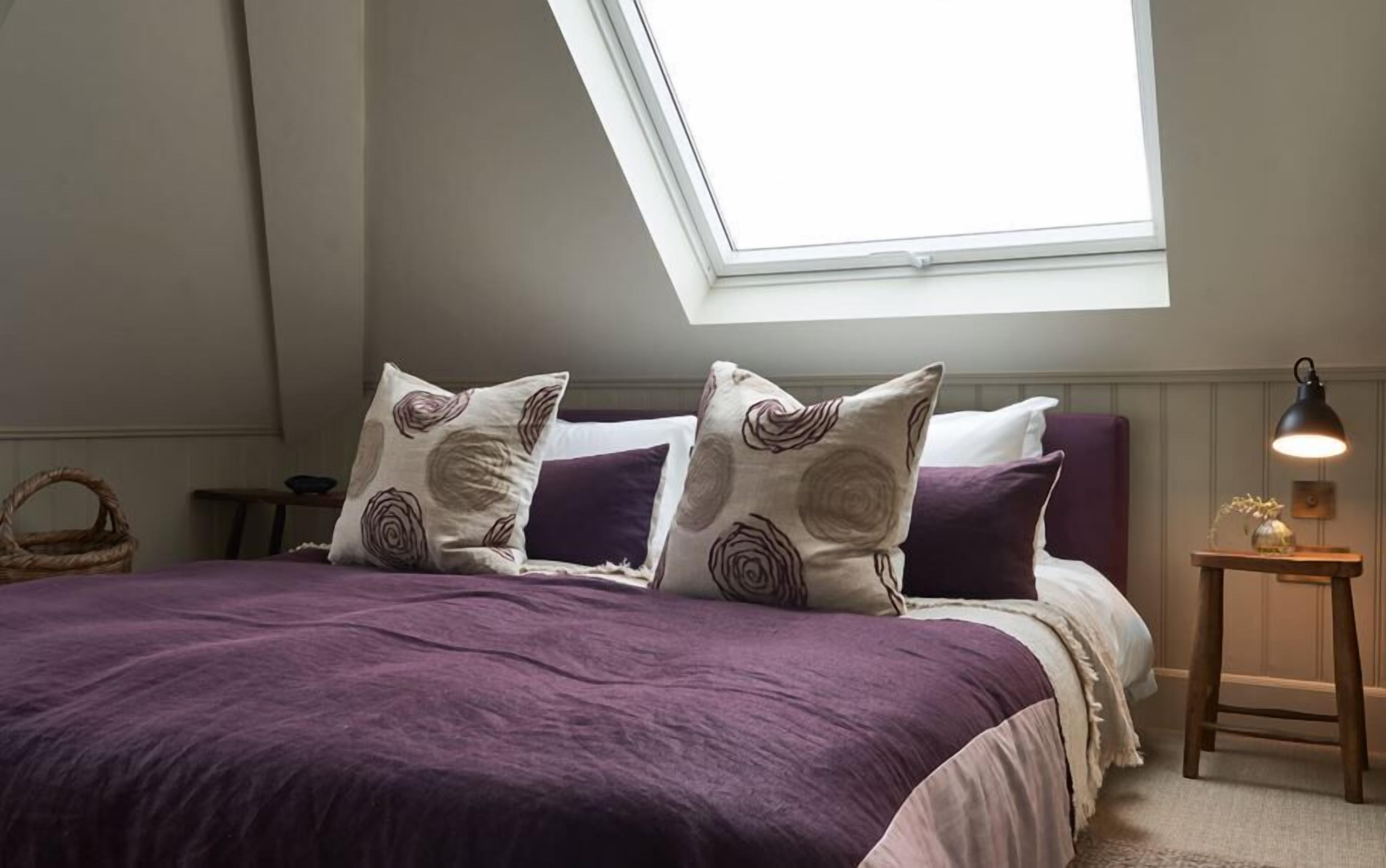 A bedroom with a skylight, a bed with purple bedding, and a bedside table with a lamp.