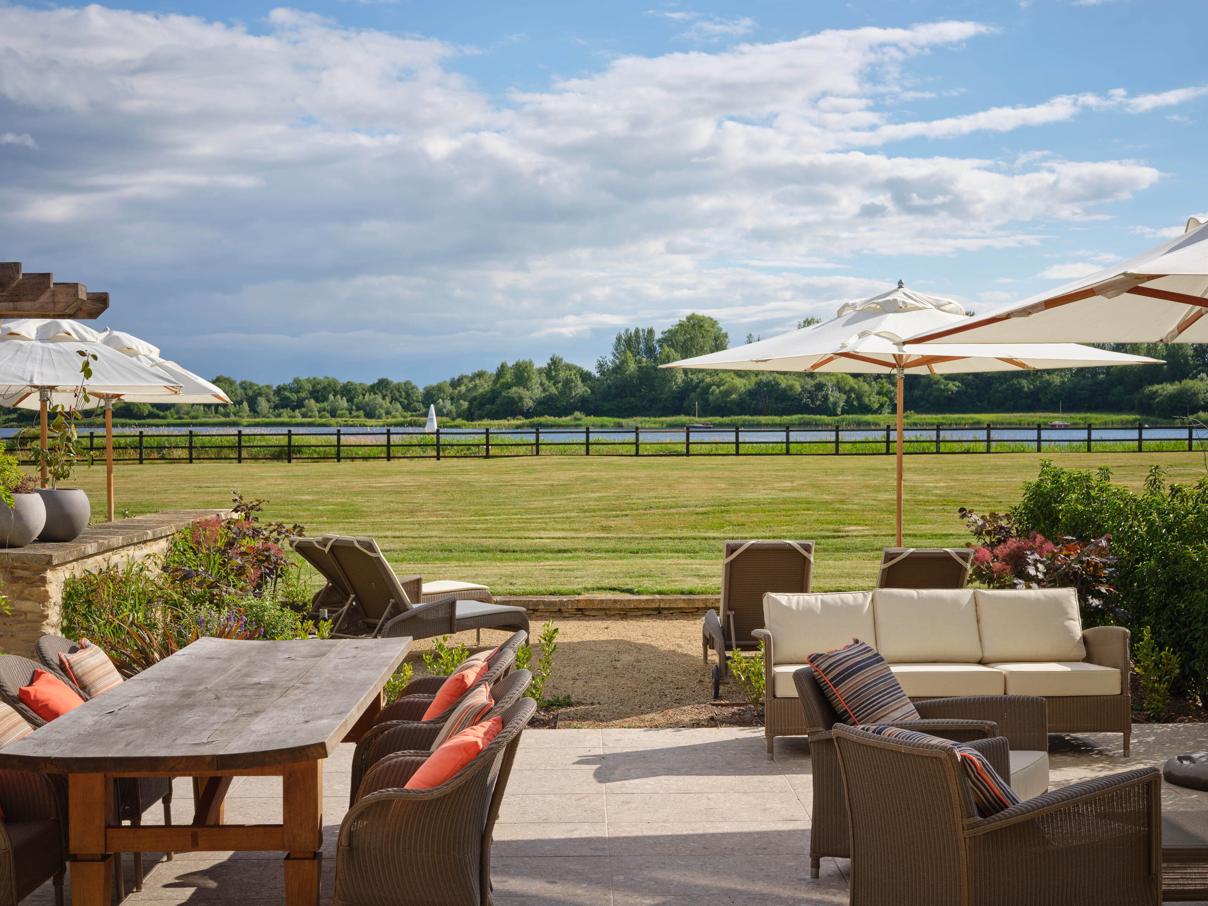An outdoor patio with wicker furniture and umbrellas, overlooking a lake and a grassy field.