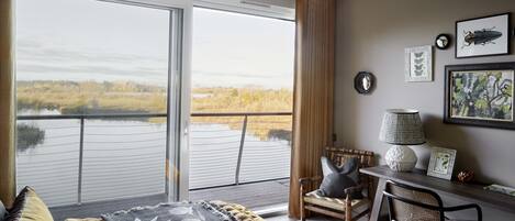 A room with a large window overlooking a body of water, a bed with a floral bedspread, a desk with a lamp, and a chair.