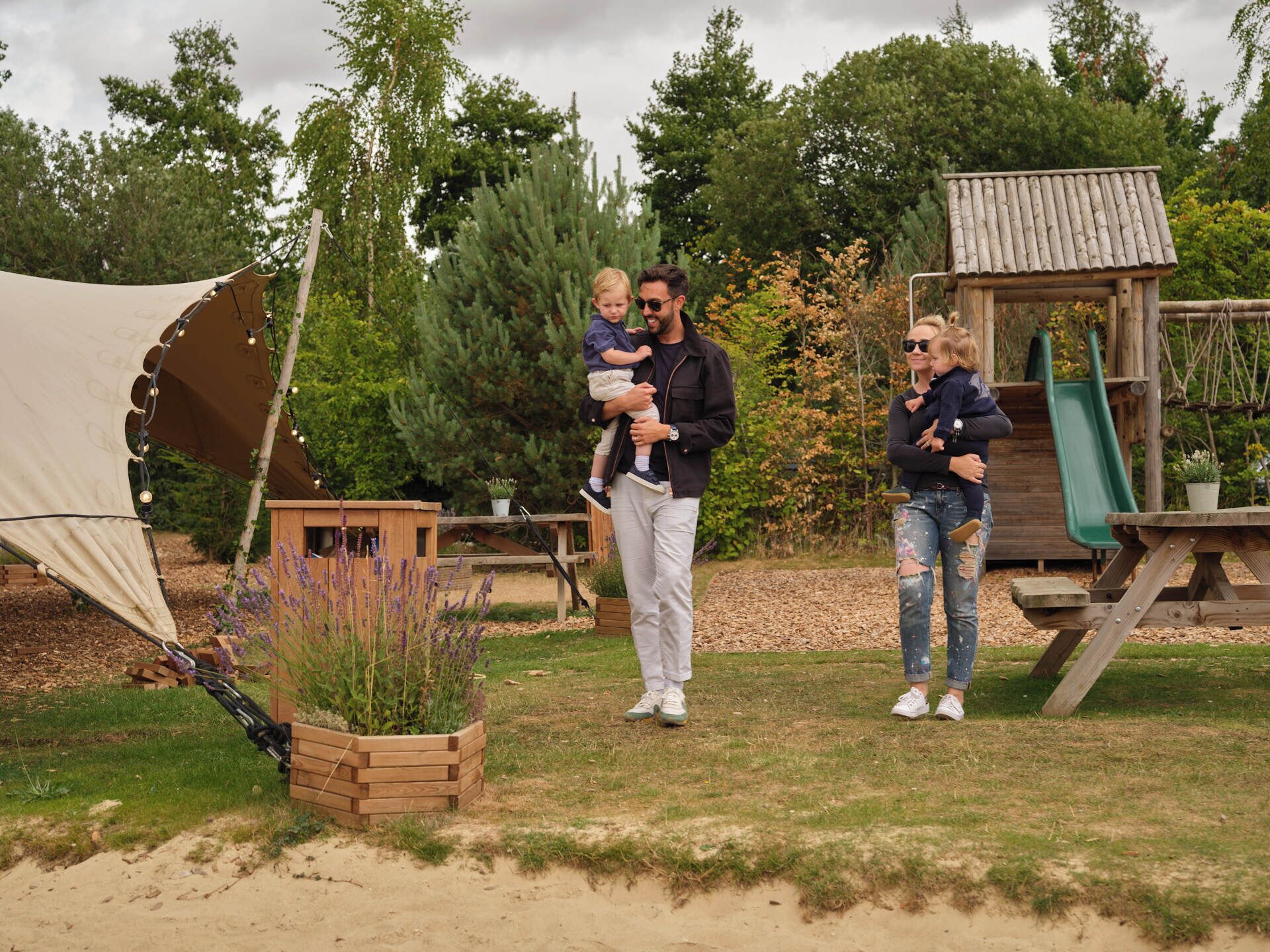 A family in a garden with a wooden play structure, a tent, and a table.