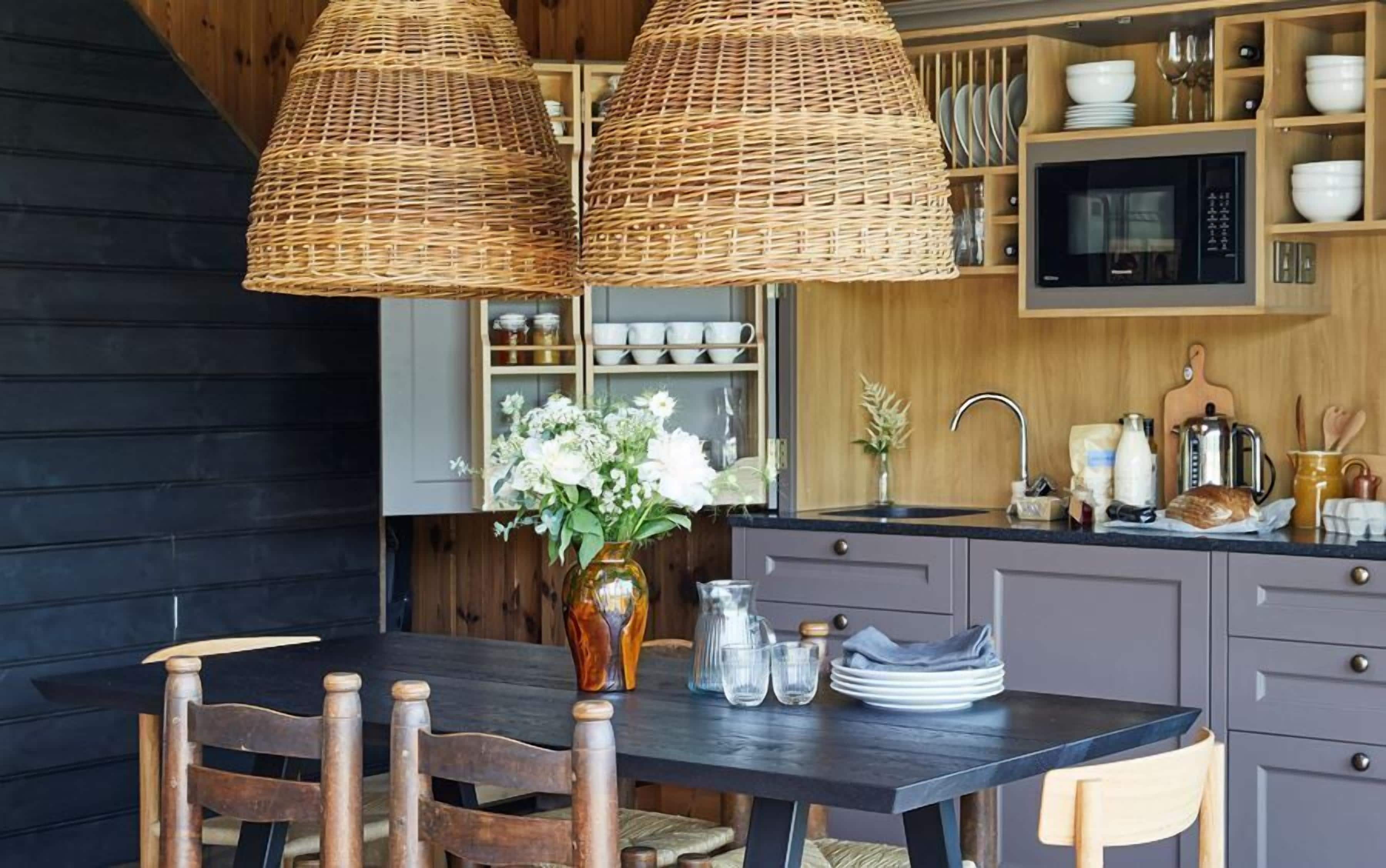 A kitchen with a dark wooden table, wicker pendant lights, and a vase with flowers.