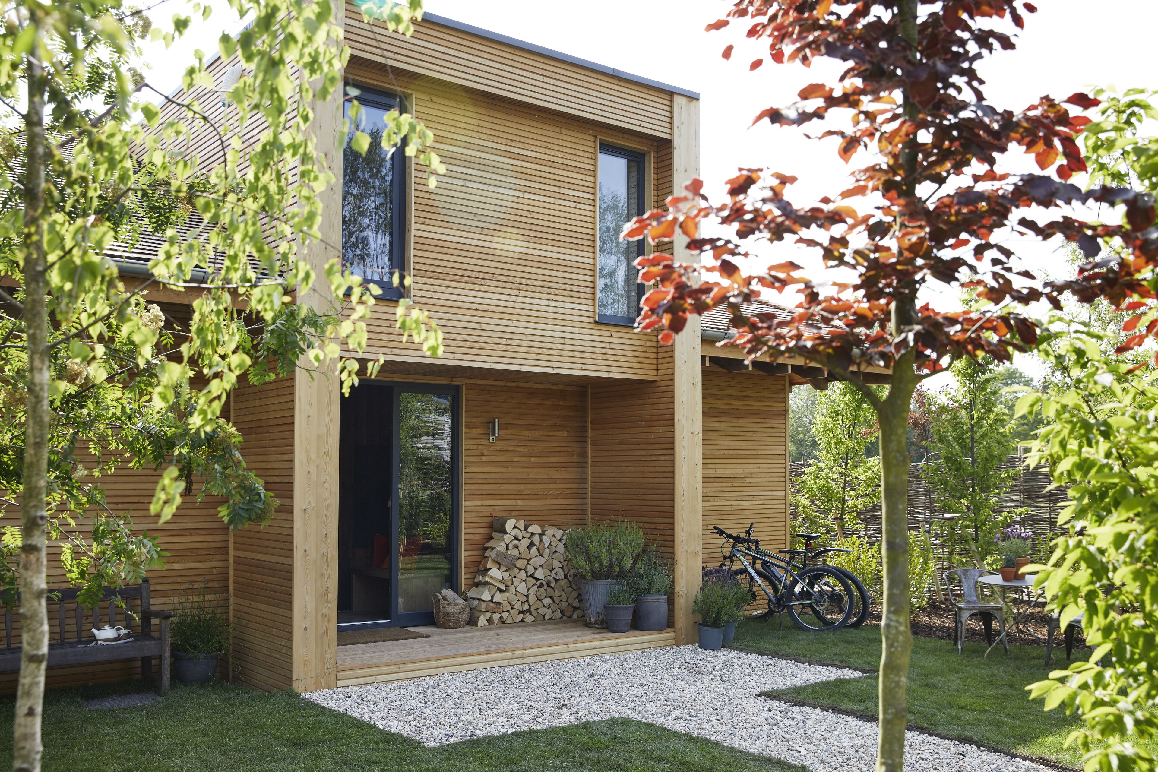 Une maison moderne à deux étages, avec une façade en bois, un porche à l’avant et un chemin de gravier menant à l’entrée.