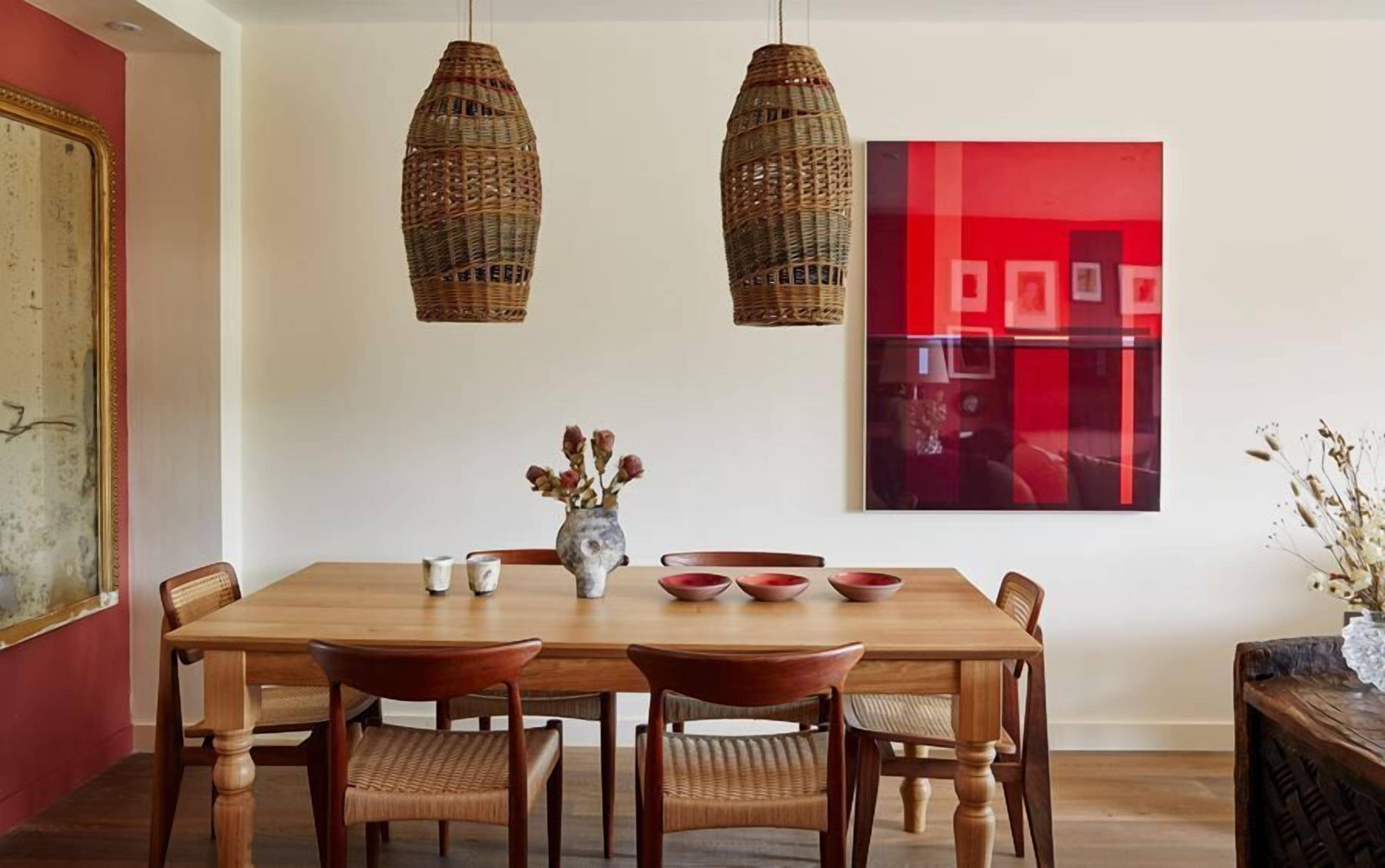 A dining room with a wooden table, six chairs, two hanging wicker chandeliers, and a vase with dried flowers.