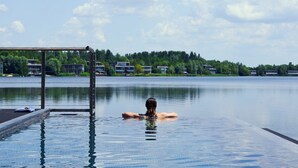 A person relaxing in a clear pool with a view of a lake and buildings in the background. - The Lakes By YOO (Lechlade)