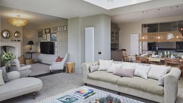 A spacious living room with a sofa, armchairs, and a coffee table. There is a fireplace, a television, and a dining area visible.