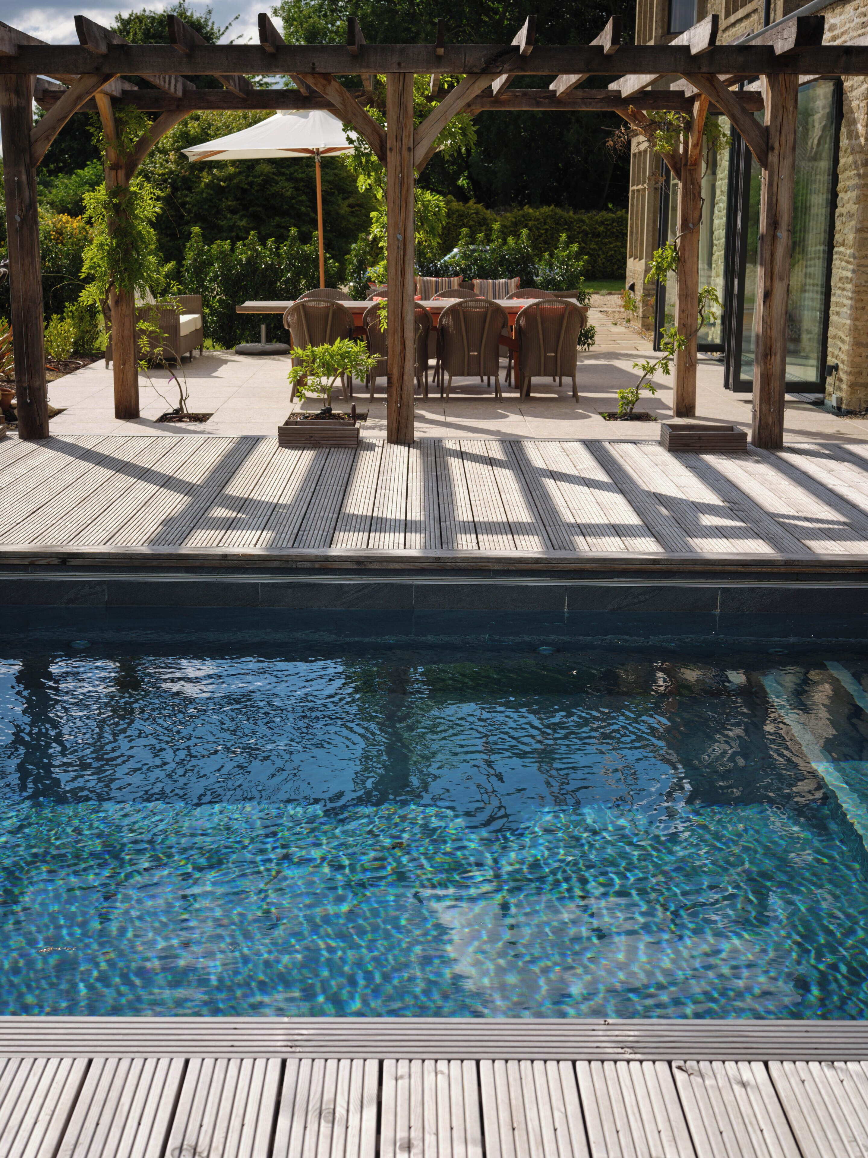 A wooden deck with a pool, outdoor furniture, and a pergola with umbrellas.