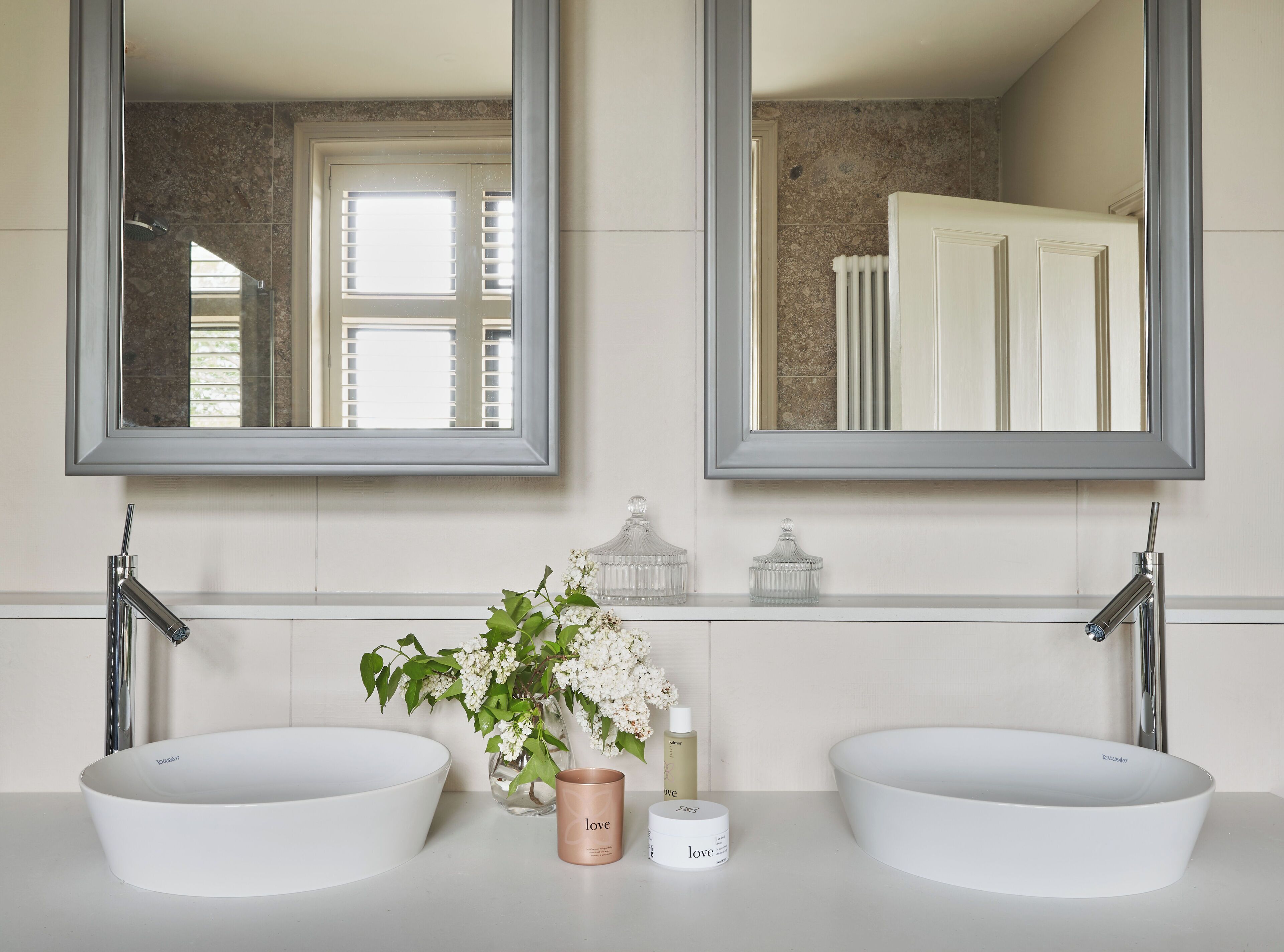 Une salle de bain avec deux lavabos blancs, un miroir et un vase rempli de fleurs.