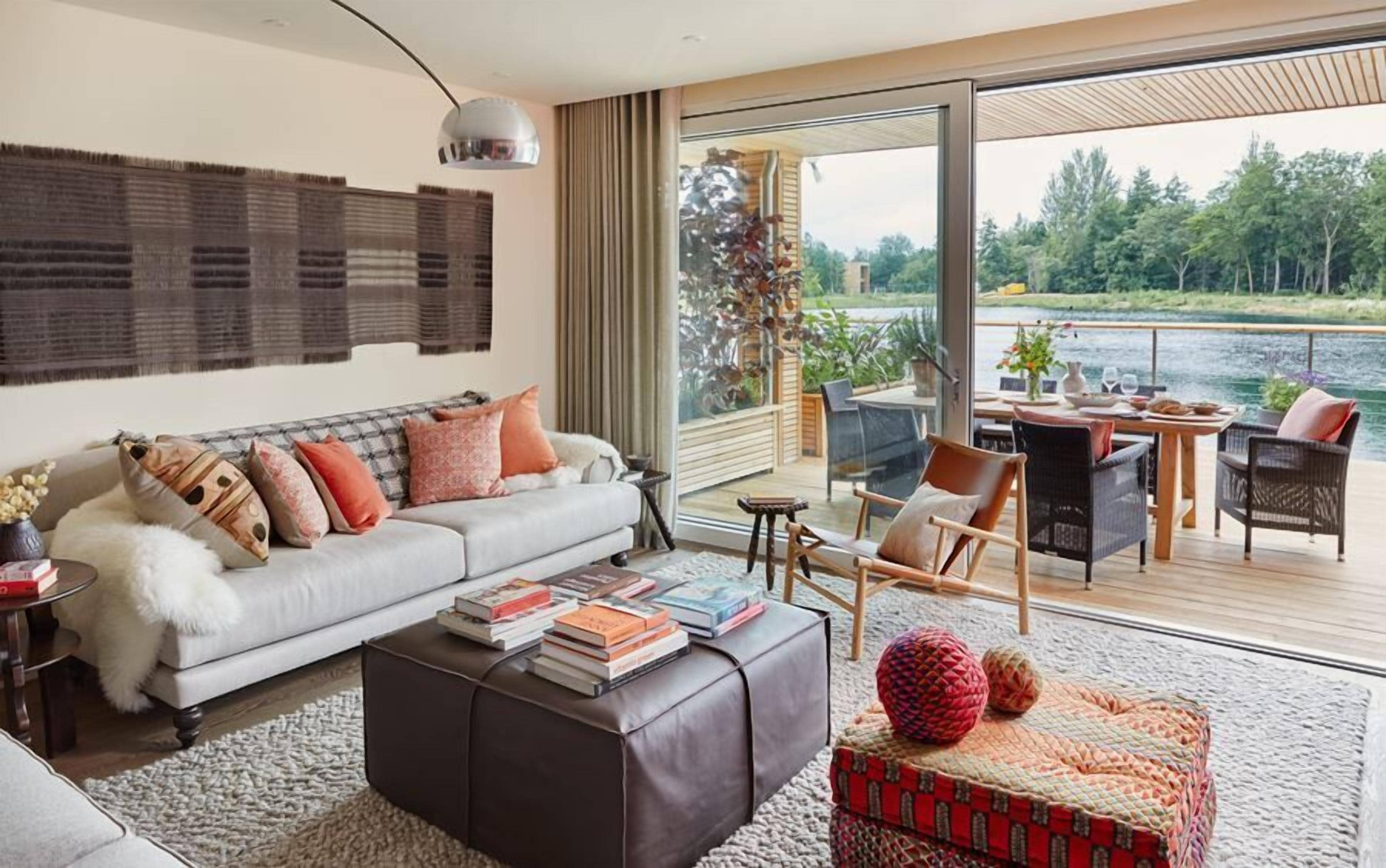 A modern living room with a sofa, coffee table, and chairs, a balcony with a dining table, and a view of a lake.