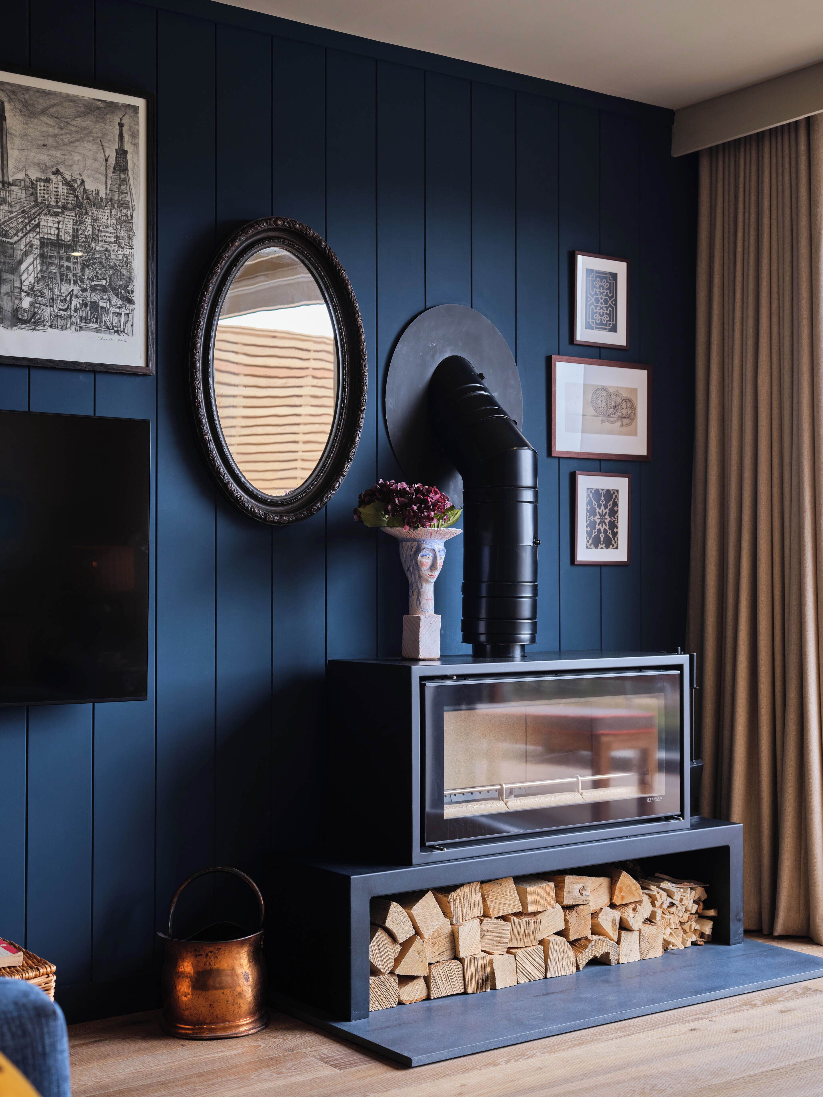 A modern living room with a dark blue wall, a fireplace with a glass door, a mounted mirror, and framed artwork.