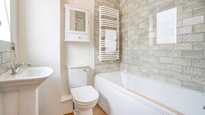 Un baño con lavamanos, inodoro y bañera blancos. Hay un toallero sobre la bañera y un armario sobre el lavamanos.