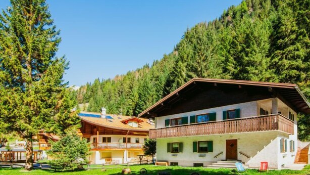 Un chalet de dos pisos con balcón, rodeado de exuberante vegetación y montañas.