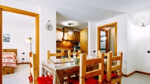 Ein Essbereich mit Holztisch und -stühlen, im Hintergrund eine Küche und im Vordergrund ein Bett mit roter Decke.