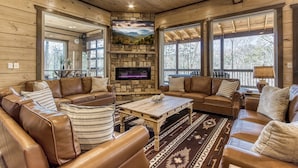 A cozy living room with leather sofas, a stone fireplace, and large windows offering a view of the outdoors. - HIBERNATION STATION - 12 Bedrooms with a pool! (Sevierville)