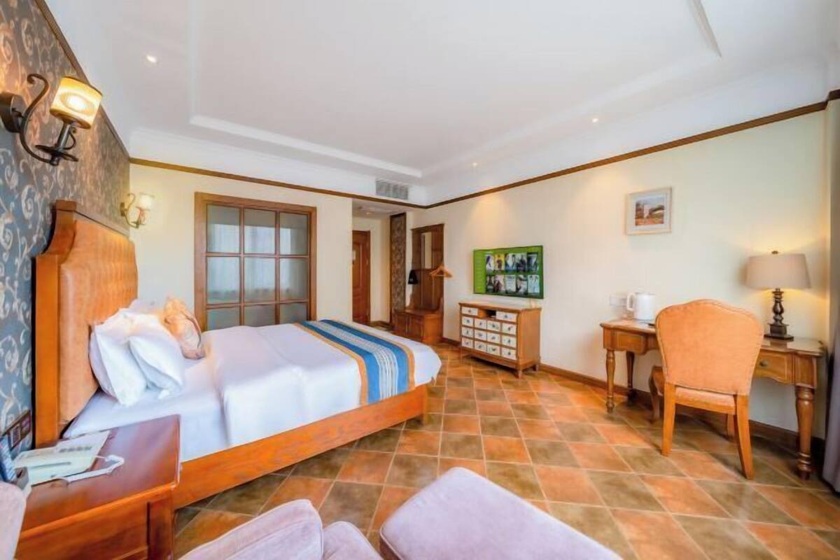 A hotel room with a large bed, a desk, a chair, a cabinet with bottles, and a painting on the wall.