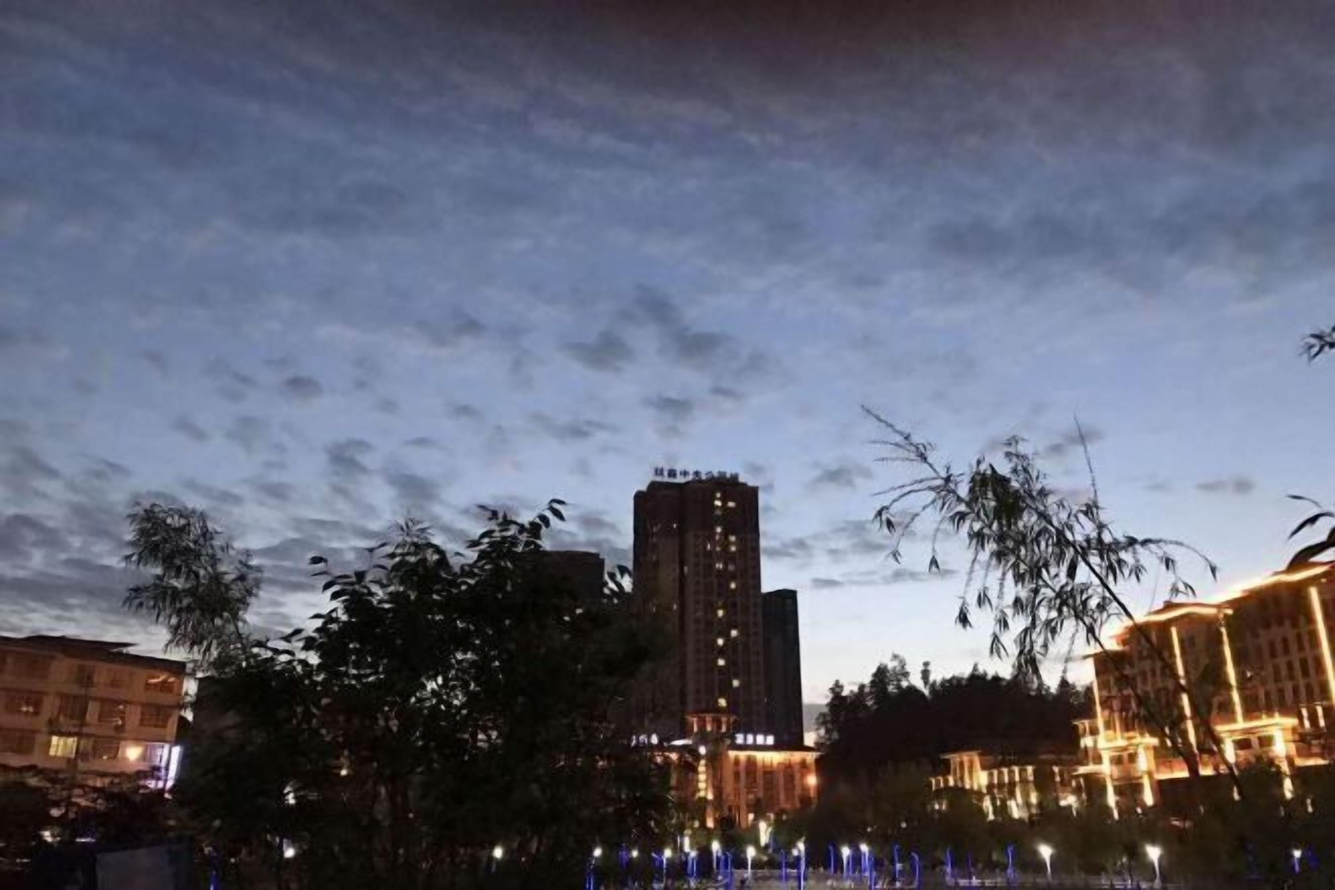 A nighttime view of a cityscape with illuminated buildings and a park area with trees and a fountain.