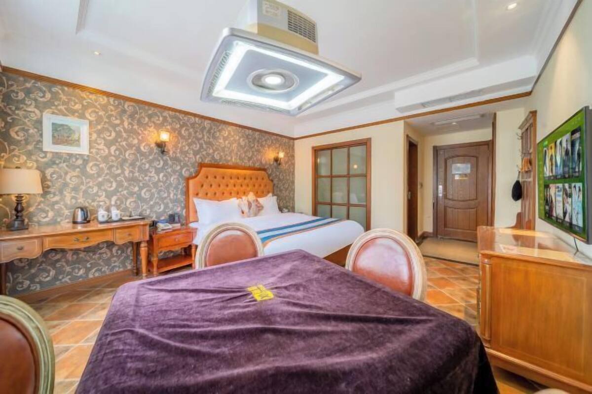 A hotel room with a bed, a desk, a chair, a table with a purple tablecloth, and a ceiling light fixture.