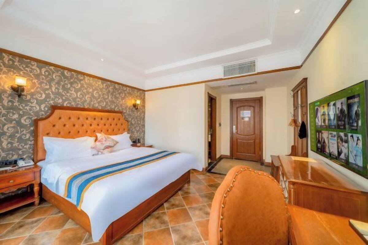 A hotel room with a large bed, a wooden headboard, a nightstand, a desk with a chair, and a wall-mounted TV.