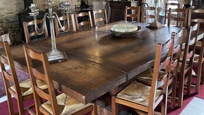 Een grote houten eettafel omringd door houten stoelen met beklede zitplaatsen, een kroonluchter en een stenen muur met een open haard.