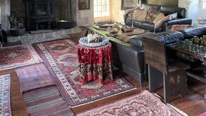 Een traditionele kamer met zichtbare houten balken, een open haard, een leren bank en vloerkleden met patronen.