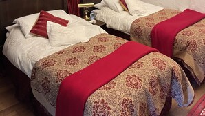 Een kamer met twee bedden, rode gordijnen, een houten hoofdbord, een klein plankje met een beeldje en een nachtkastje met een lamp en een waterfles.