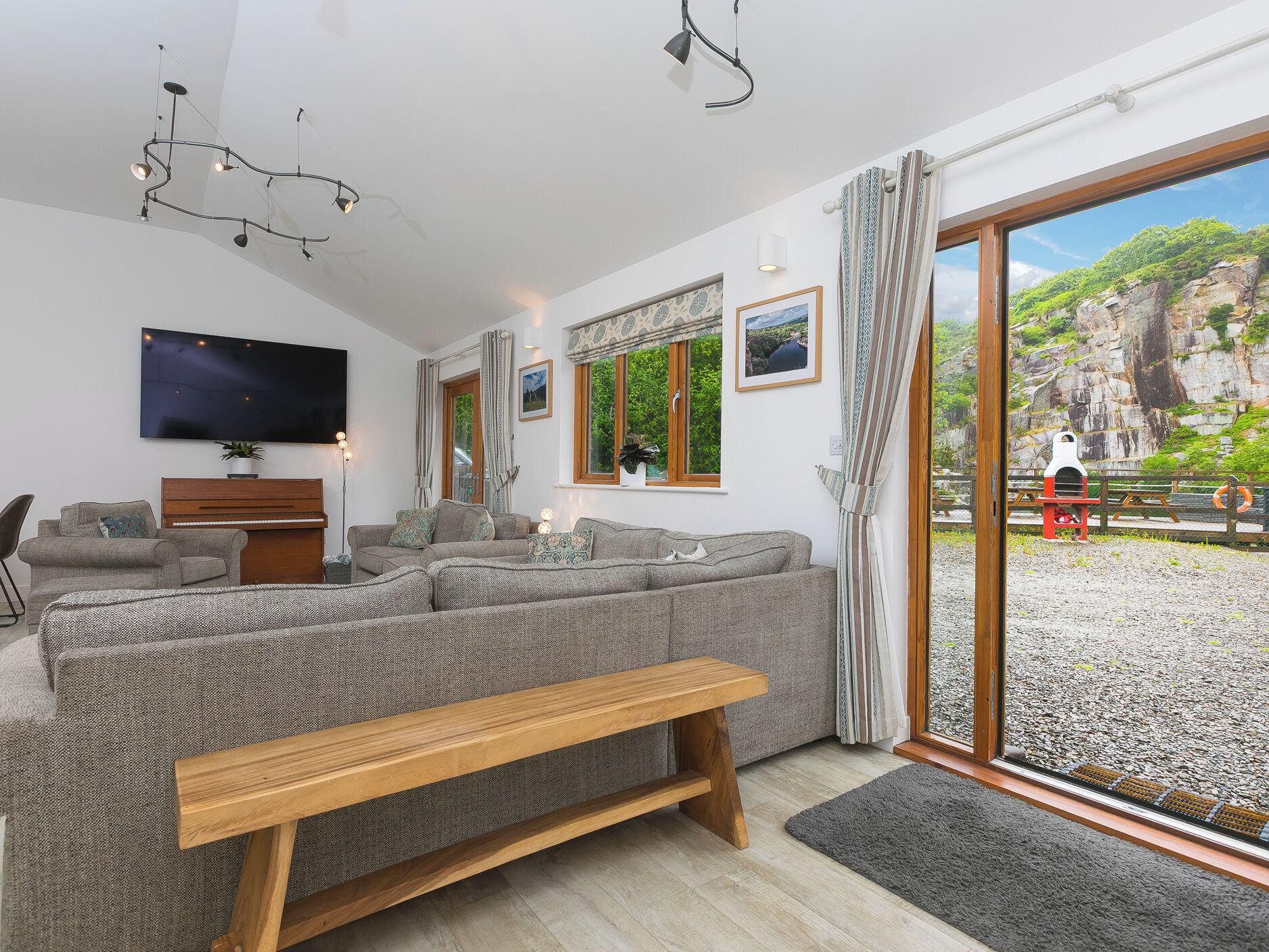 A modern living room with a sofa, a wooden bench, a flat-screen TV, and a view of a rocky landscape through large windows.