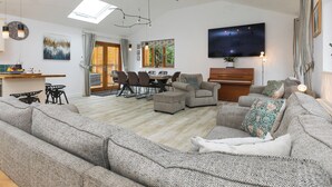 Amplia sala de estar con un sofá grande, un comedor con sillas, un piano y un televisor montado en la pared.