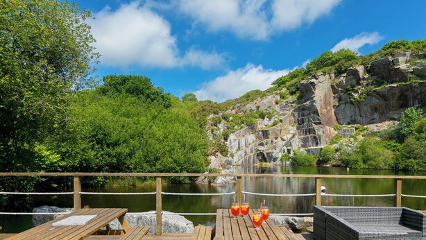 Terraza de madera con mesas de picnic y mobiliario de mimbre, con vistas a un lago y un acantilado rocoso al fondo.