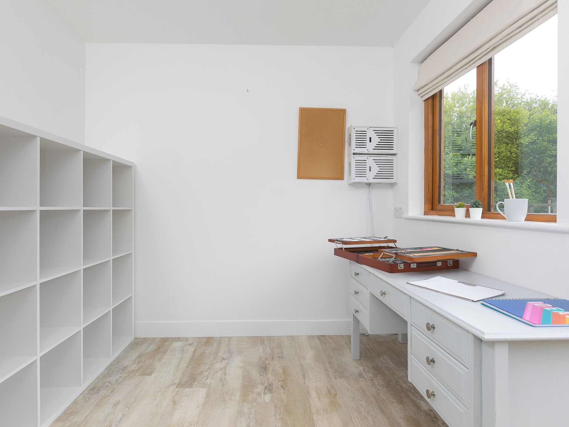 Una habitación con un escritorio blanco, un corcho y un ventanal con vistas a la vegetación.