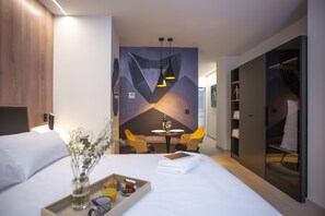 Ein modernes Hotelzimmer mit Bett, Esstisch mit Stühlen, einer Minibar und einem Kleiderschrank.