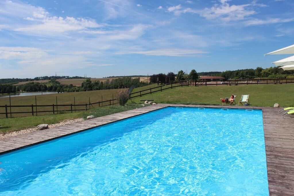 A rectangular swimming pool with a wooden deck, surrounded by a wooden fence, and a grassy area with a few chairs and a person.