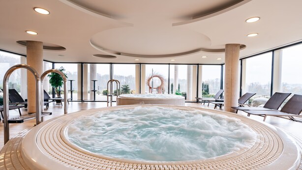 A large indoor hot tub with a view of a cityscape through large windows. - K-Triumf resort (Velichovky)