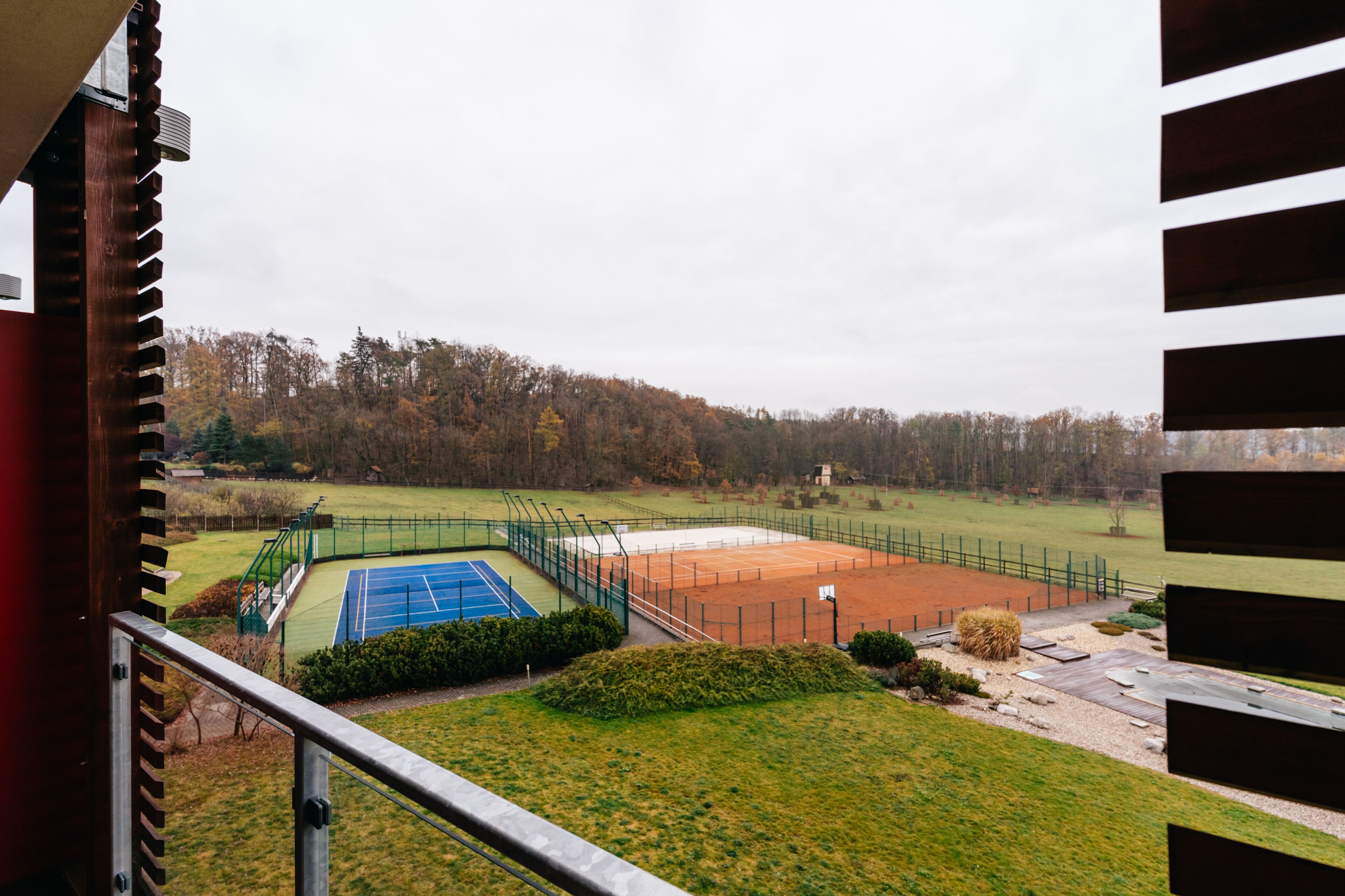 A tennis court with a green fence, a basketball court, and a grassy area.