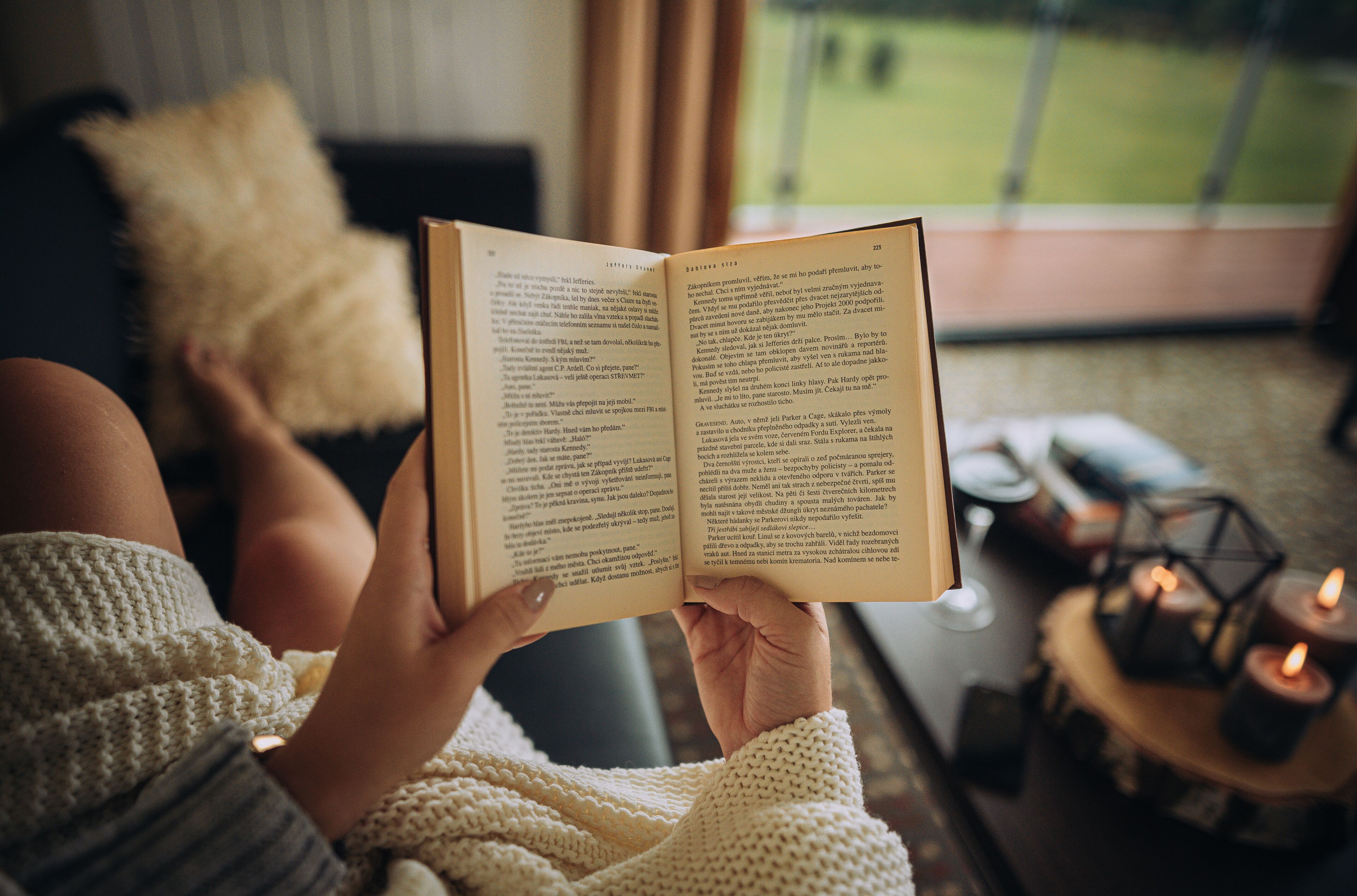 A person reading a book in a cozy room with a view of a green landscape through a window.