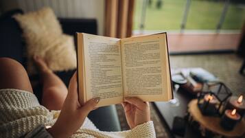 A person reading a book in a cozy room with a view of a green landscape through a window.