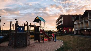 A playground with climbing structures and a slide, surrounded by a sandy area and a building with balconies.