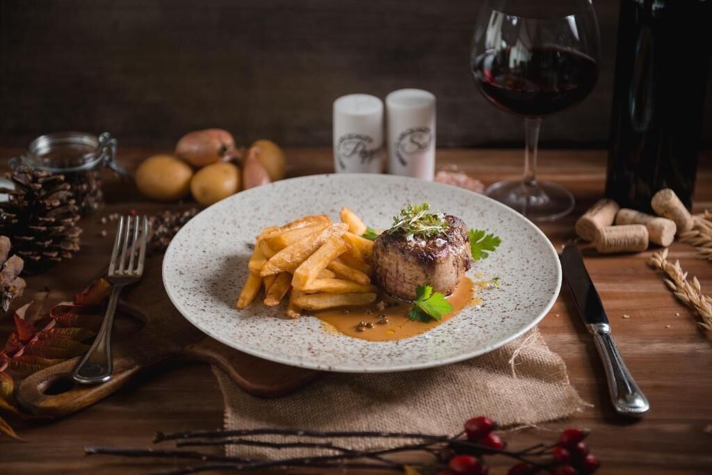 En tallerken mad med steak, pommes frites og sauce, ledsaget af et glas rødvin, salt- og peberkværke, og et rustikt miljø med trædetaljer.
