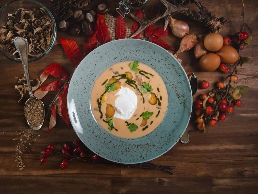 A bowl of soup with a dollop of cream and garnishes, surrounded by autumn leaves and berries on a wooden table.