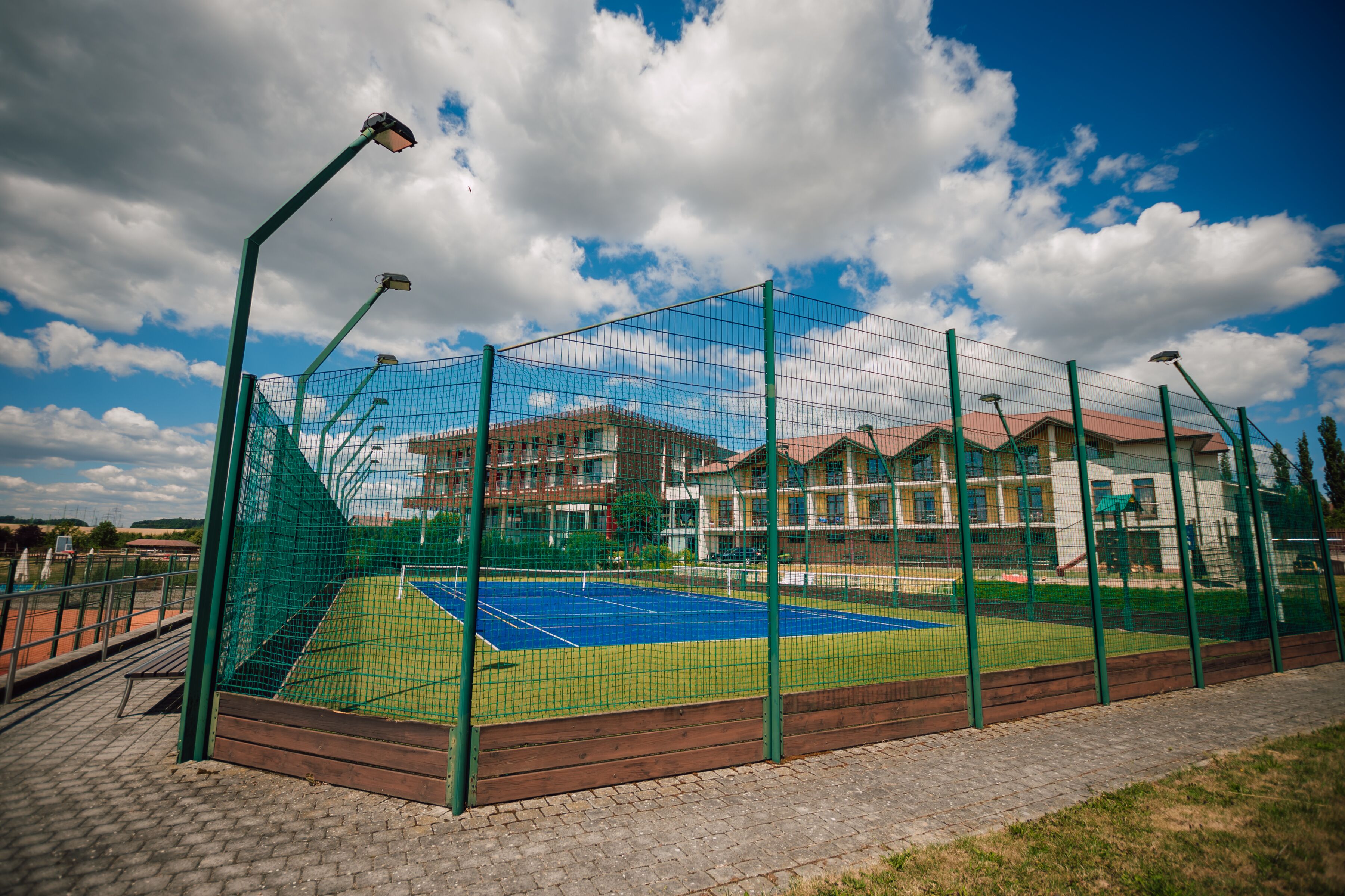 A tennis court with a net, surrounded by a green fence, and buildings in the background.