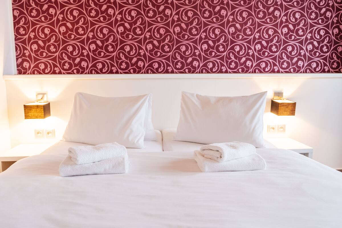 A neatly made bed with white linens and two pillows, a red patterned wallpaper above, and two bedside lamps on either side.