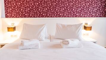 A neatly made bed with white linens and two pillows, a red patterned wallpaper above, and two bedside lamps on either side.