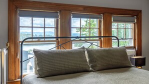 Una cama con estructura metálica y dos almohadas, un marco de ventana de madera, y vista a un lago y montañas.