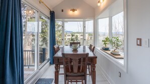 Un comedor con una mesa grande y sillas, ventanas amplias con vistas a árboles y edificios, y un techo con una lámpara.