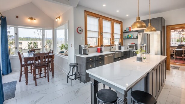 Cocina moderna con una isla grande, muebles oscuros y electrodomésticos de acero inoxidable. Cuenta con un comedor con mesa y sillas, y amplias ventanas que ofrecen luz natural.